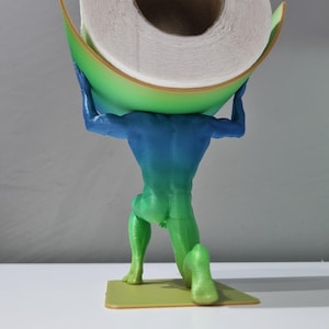 May include: A green, blue, and yellow 3D printed figurine of a person holding a roll of toilet paper. The figurine is kneeling on a small, yellow base.