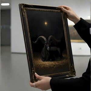 May include: A framed artwork depicting a black goat with red eyes, resting on straw against a dark backdrop. A golden star shines above the goat. The ornate frame has a gold and black finish.
