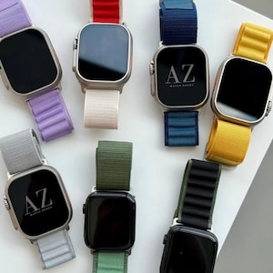 May include: A collection of smartwatches with various colored bands, including purple, red, white, blue, yellow, gray, green, and black. Each watch face displays the letters "AZ" and the words "WATCH BANDS". The watches have a square screen and a silver-colored case.