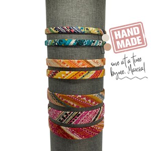 May include: A set of six colorful patterned headbands displayed on a gray stand. The headbands are made of fabric with different geometric patterns in shades of blue, orange, yellow, pink, and white. The text "HAND MADE one at a time by me. Marcia!" is visible in the image.