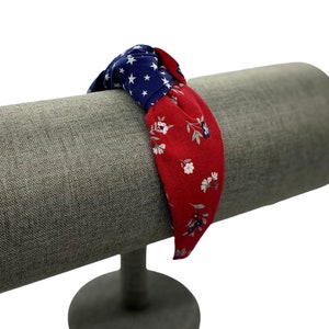 May include: Red and blue fabric headband with a knotted design and a floral pattern. The headband is displayed on a gray fabric covered stand.