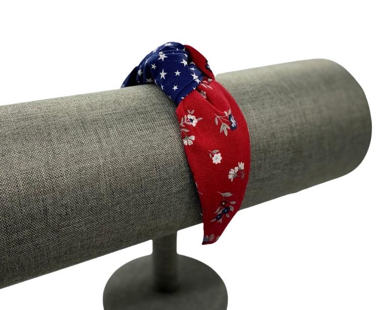 May include: Red and blue fabric headband with a knotted design and a floral pattern. The headband is displayed on a gray fabric covered stand.