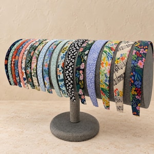 May include: A collection of fifteen fabric-covered headbands displayed on a gray cylindrical stand. The headbands feature various patterns including floral prints in red, blue, green, and yellow, polka dots, and a musical staff print on a cream background.