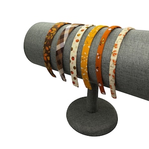 May include: A collection of seven fabric headbands with autumn-themed patterns. The headbands are displayed on a gray cylindrical stand. Patterns include leaves, plaid, and floral designs in shades of brown, orange, and yellow.