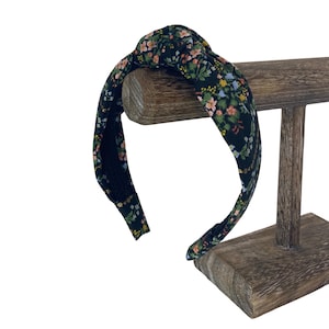 May include: A black fabric headband with a knotted top, adorned with a floral pattern in shades of pink, yellow, and green. The headband is displayed on a wooden stand, showcasing its design and style. This accessory is perfect for adding a touch of elegance to any hairstyle.