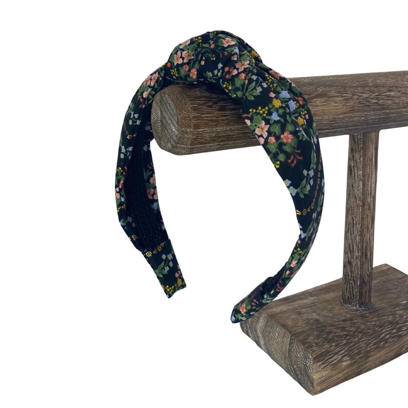 May include: A black fabric headband with a knotted top, adorned with a floral pattern in shades of pink, yellow, and green. The headband is displayed on a wooden stand, showcasing its design and style. This accessory is perfect for adding a touch of elegance to any hairstyle.