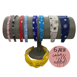 May include: Six colorful headbands with a bandana print. The headbands are displayed on a headband stand. The headbands are in the colors pink, black, red, light blue, dark blue, and white.  The text "6 NEW colors added!" is displayed on a pink circle.
