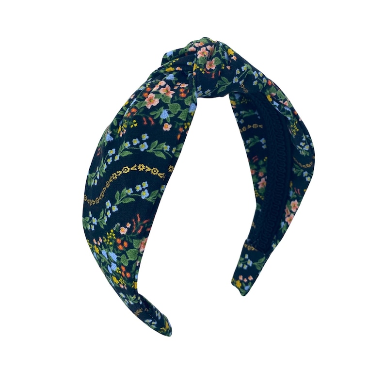 May include: A dark blue fabric headband with a knotted top. The headband is decorated with a floral pattern in shades of pink, yellow, and green. The headband is a stylish hair accessory.