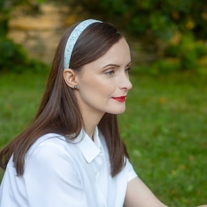 Peut inclure: Un bandeau bleu clair orn&eacute; d'un motif floral blanc. Le bandeau est port&eacute; par une personne aux longs cheveux bruns, au rouge &agrave; l&egrave;vres rouge et &agrave; une chemise blanche &agrave; col. L'arri&egrave;re-plan est un feuillage vert flou.