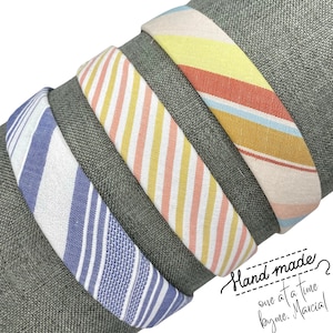 May include: Four fabric headbands with striped patterns in blue, pink, yellow, and orange. The headbands are stacked on top of each other. The text "Hand made? One at a time by me. Marcia!" is written in cursive on a dark green background.