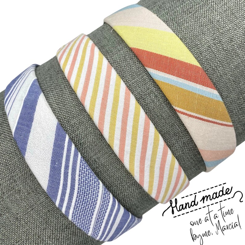 May include: Four fabric headbands with striped patterns in blue, pink, yellow, and orange. The headbands are stacked on top of each other. The text "Hand made? One at a time by me. Marcia!" is written in cursive on a dark green background.