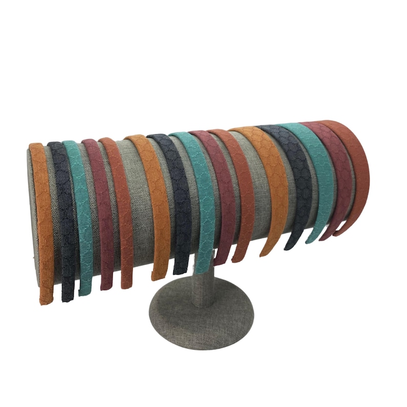May include: A collection of fourteen thin headbands arranged on a cylindrical display stand. Each headband features a textured, honeycomb-like pattern and comes in a variety of colors including orange, dark blue, teal, maroon, and rust brown.