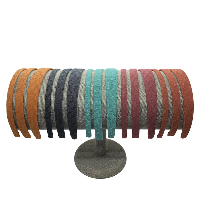 May include: A collection of fourteen thin headbands arranged in a row on a display stand. The headbands feature a textured, repeating geometric pattern and come in a variety of colors including orange, dark gray, teal, maroon, and terracotta.