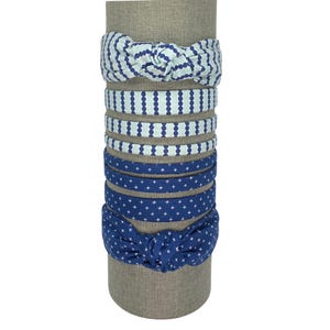 May include: A collection of fabric headbands in various patterns and colors, displayed on a cylindrical stand. The headbands feature designs like polka dots, cross patterns, and knotted bows. Colors include shades of blue and white.