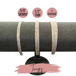 May include: Three white fabric headbands with a twig and leaf pattern in shades of orange, brown, and cream. The headbands are displayed on a gray fabric covered stand. The headbands are labeled 3/8" Skinny, 1/2" Thin, and 1" Average.