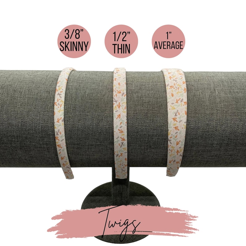 May include: Three white fabric headbands with a twig and leaf pattern in shades of orange, brown, and cream. The headbands are displayed on a gray fabric covered stand. The headbands are labeled 3/8" Skinny, 1/2" Thin, and 1" Average.