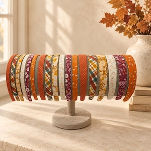 Op de afbeelding: Een collectie met stof beklede haarbanden op een horizontale standaard. De haarbanden hebben diverse herfstachtige patronen, waaronder bloemenprints, ruiten, stippen en effen kleuren in tinten oranje, geel, bordeauxrood en crème.