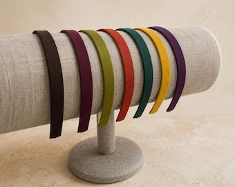 May include: A set of seven thin, fabric-covered headbands arranged in a row. The headbands feature a smooth, matte texture and come in a variety of solid colors including dark brown, burgundy, olive green, burnt orange, dark teal, mustard yellow, and deep purple.
