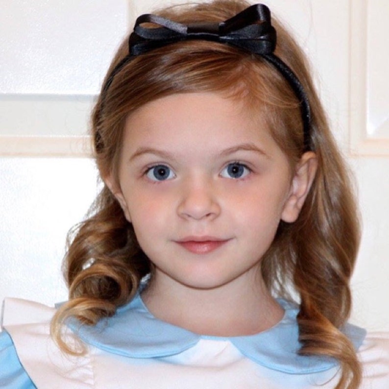 May include: A young girl with long brown hair wearing a blue and white dress with a Peter Pan collar and a black satin bow headband.