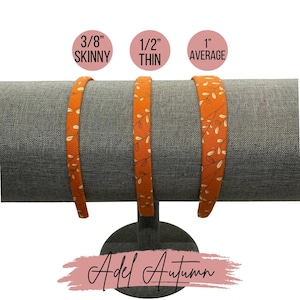 May include: Three orange fabric headbands with a white floral pattern, displayed on a stand. The headbands are labeled "3/8" Skinny", "1/2" Thin", and "1" Average".