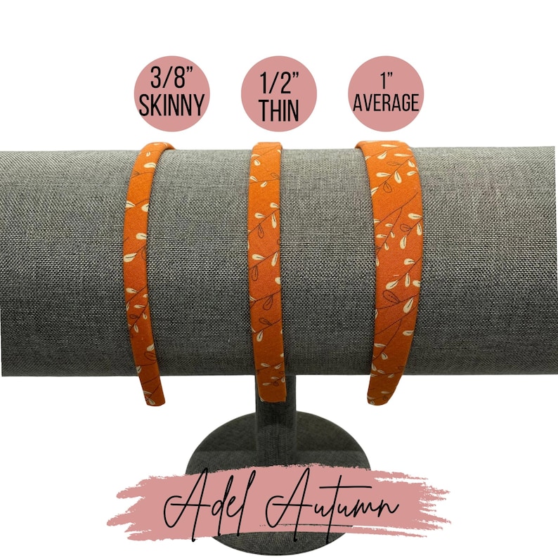 May include: Three orange fabric headbands with a white floral pattern, displayed on a stand. The headbands are labeled "3/8" Skinny", "1/2" Thin", and "1" Average".