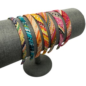 May include: A set of eight colorful patterned headbands displayed on a gray headband stand. The headbands feature various geometric and floral designs in shades of pink, blue, yellow, and orange.