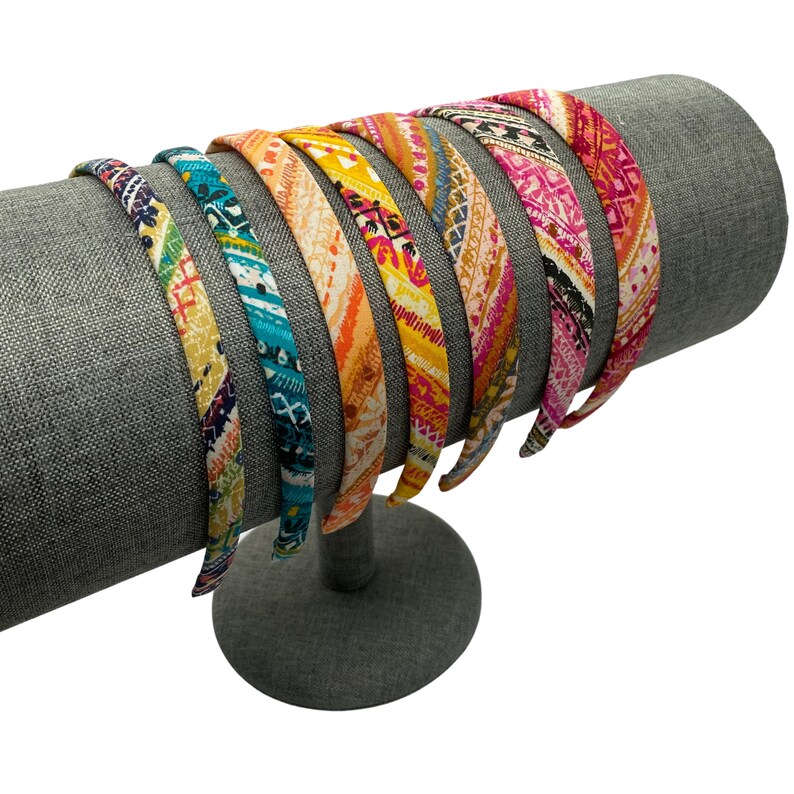 May include: A set of eight colorful patterned headbands displayed on a gray headband stand. The headbands feature various geometric and floral designs in shades of pink, blue, yellow, and orange.