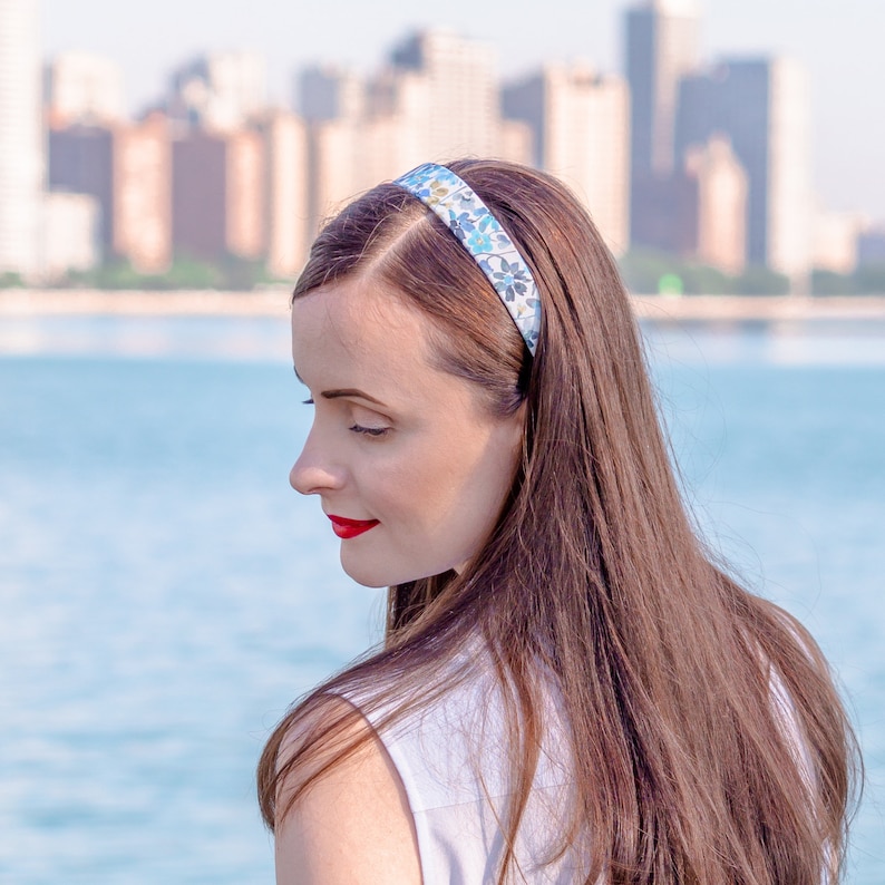 May include: A floral headband with blue and white flowers is worn by a person with long brown hair. The headband is approximately 1 inch wide. The person is wearing a white top and red lipstick. The background features a body of water and a city skyline.