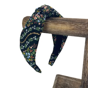 May include: A black fabric headband with a knotted top, adorned with a colorful floral pattern. The flowers are in shades of pink, yellow, blue, and green. The headband is displayed on a wooden stand.