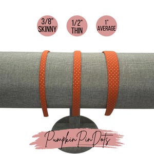 May include: Three orange headbands with white polka dots on a gray fabric stand. The headbands are labeled 3/8" Skinny, 1/2" Thin, and 1" Average. The text "Pumpkin Pin Dots" is written in pink on a white background.
