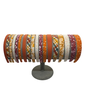 May include: A display of 18 headbands with various fall-themed patterns in shades of orange, brown, and white. The headbands are arranged in a circle on a gray stand.