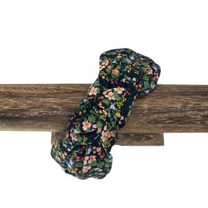 May include: A floral print headband with a dark background and a pattern of pink, yellow, and blue flowers. The headband is knotted and rests on a wooden surface.