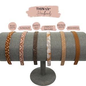 Op de afbeelding: Zes verschillende gepatroneerde dunne 1/2 inch hoofdbanden die op een hoofdbandenstandaard worden getoond. De hoofdbanden zijn gelabeld met "Rust Dots", "Sundazes", "Sweet Sky", "Unruly Nature", "Bronze Dots" en "Mishmesh".