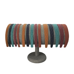May include: A collection of fourteen thin headbands arranged in a row, featuring a textured, repeating geometric pattern. The headbands come in a variety of colors including shades of orange, dark blue, teal, and maroon, all displayed on a stand.
