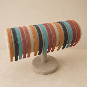 May include: A collection of thin headbands arranged on a cylindrical display stand. The headbands feature a textured surface and come in a variety of colors including orange, dark gray, teal, maroon, and rust, all lined up side by side.