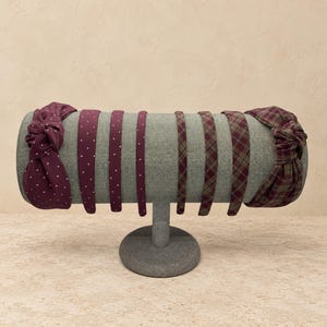 May include: A collection of eight headbands displayed on a cylindrical stand. The set includes knotted and slim styles in deep burgundy with white polka dots, solid burgundy, and a burgundy and olive green plaid fabric pattern.