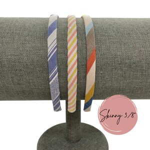 May include: Three fabric headbands on a gray display stand. The headbands are striped in blue and white, pink and yellow, and red and brown. The text "Skinny 3/8" is written on a pink circle.