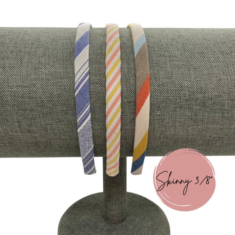 May include: Three fabric headbands on a gray display stand. The headbands are striped in blue and white, pink and yellow, and red and brown. The text "Skinny 3/8" is written on a pink circle.