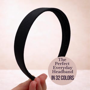 May include: A black headband with a smooth, matte finish. The image text reads "The Perfect Everyday Headband IN 32 COLOURS". The headband is held by a hand against a soft, neutral background.
