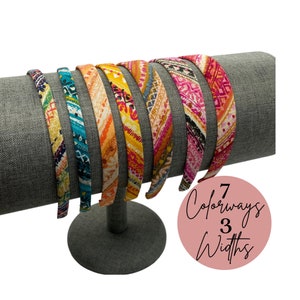 May include: Seven colorful patterned headbands displayed on a gray headband stand. The headbands are in various widths and feature a mix of pink, yellow, blue, and orange colors.