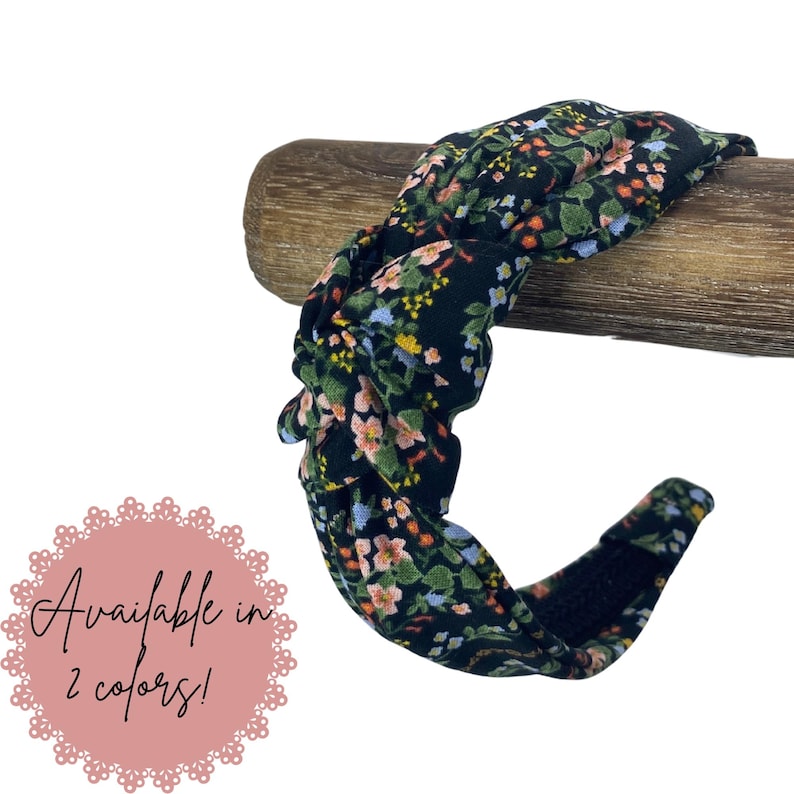 May include: A black floral headband with a knotted design. The headband features a pattern of small pink, blue, and yellow flowers with green leaves. The text "Available in 2 colors!" is displayed on a pink background.