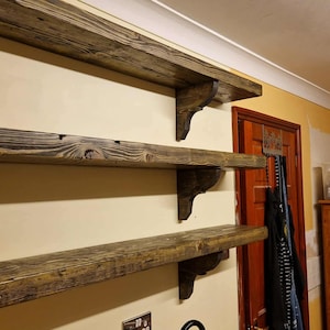 May include: Three rustic wooden shelves mounted on a beige wall. The shelves are made of weathered wood with visible grain and knots, supported by decorative corbels. The shelves are a warm brown color.