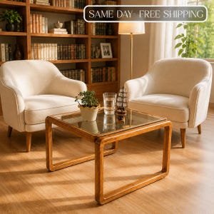May include: Two cream-coloured upholstered armchairs with rounded backs and wooden legs positioned on either side of a square wooden coffee table with a glass top. A small potted plant, a clear glass, and a model ship sit on the table surface.