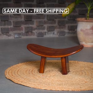 Puede incluir: Un taburete de madera marrón oscuro con asiento curvo y tres patas, colocado sobre una alfombra redonda de yute. El diseño del taburete es simple y elegante. El fondo incluye una pared de ladrillo y una planta en maceta de terracota. El texto "SAME DAY - FREE SHIPPING" es visible.