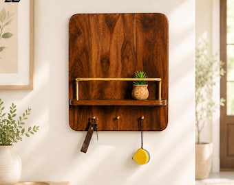 May include: A rectangular wall-mounted organiser made of dark brown wood with a rounded top edge. It features a horizontal gold metal rail, a small shelf holding a potted succulent, and three gold hooks. A leather keychain and a yellow tape measure hang from the hooks.