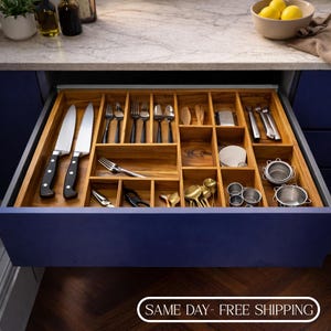 Puede incluir: Un organizador de cajón de cocina de madera lleno de varios utensilios. El cajón contiene cuchillos, tenedores, cucharas y otras herramientas de cocina. El organizador es de madera y tiene múltiples compartimentos para almacenamiento. El cajón es azul y está abierto.