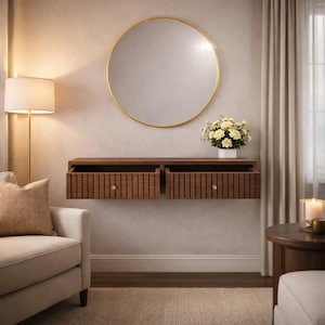 May include: A walnut-effect floating console with two open drawers, ribbed detailing, and gold knobs. A large circular mirror with a gold frame is mounted above. A vase of white flowers sits on the console. A floor lamp and armchair are in the foreground.