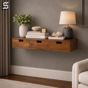 May include: A wall-mounted wooden floating shelf featuring three drawers with rectangular cut-out handles. The shelf surface holds a white vase with a green plant, a bowl of small items, stacked books labelled Architectural Digest and Interior Design.