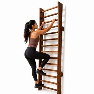 May include: A wooden stall bar climbing ladder with horizontal rungs. The ladder is made of dark wood and is mounted against a white wall. The person is climbing the ladder, wearing black leggings and a brown tank top.