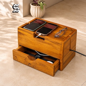 May include: A rectangular wooden charging station with a natural finish, featuring a top surface for two smartphones and spectacles, and a pull-out drawer containing a black wristwatch. A black power cable extends from the side of the unit.